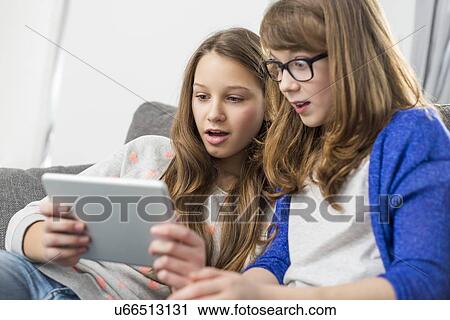 Stock Image - Shocked sisters using digital tablet on sofa at home. Fotosearch