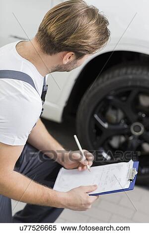 Stock Photography - Side view of automobile mechanic writing on clipboard while examining car's wheel in workshop. Fotosearch