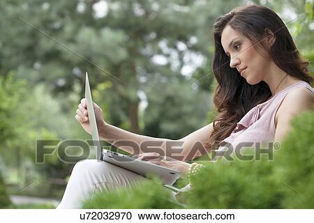 Stock Image - Side view of beautiful young woman using laptop in park. Fotosearch