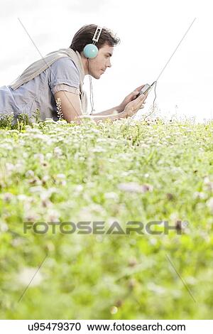Stock Image - Side view of man listening to music on MP3 player using headphones while lying in park against clear sky. Fotosearch