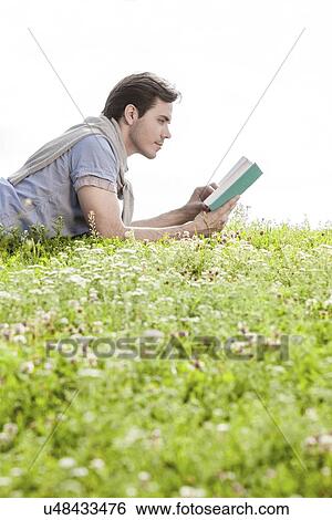 Stock Photograph - Side view of man reading book while lying on grass against clear sky. Fotosearch