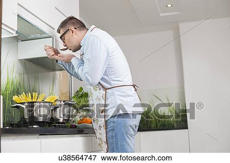Side view of man tasting food while cooking in kitchen View Large Photo Image Stock Photo - Side view of man tasting food while cooking in kitchen. Fotosearch