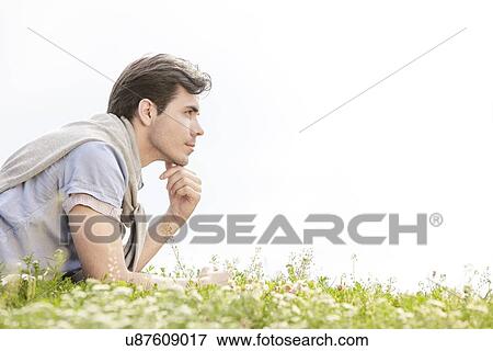 Stock Photo - Side view of thoughtful young man lying on grass against clear sky. Fotosearch