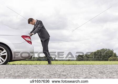 Stock Photograph - Side view of tired young businessman pushing car at countryside. Fotosearch