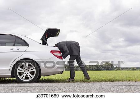 Side view of young businessman looking in trunk of car at countryside View Large Photo Image Stock Photo - Side view of young businessman looking in trunk of car at countryside. Fotosearch