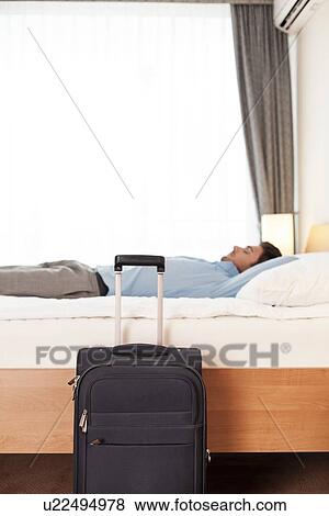 Stock Photo - Side view of young businessman sleeping in bed by luggage at hotel. Fotosearch