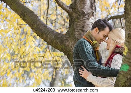 Stock Image - Side view of young couple hugging near autumn tree in park. Fotosearch