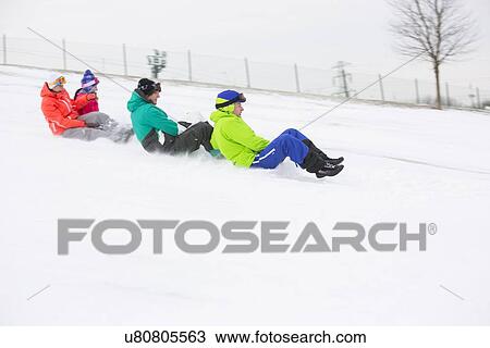 Stock Image - Side view of young friends sledging on snow covered slope. Fotosearch