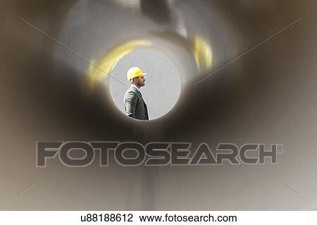 Stock Image - Side view of young male architect through large pipe at construction site. Fotosearch