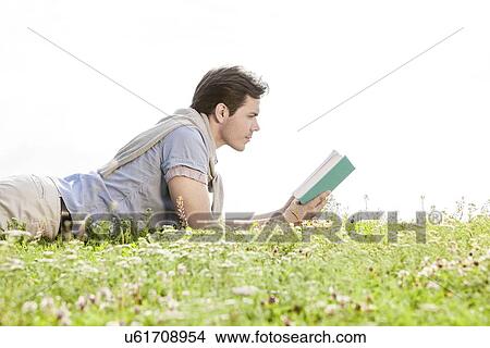 Side view of young man reading book while lying on grass against clear sky View Large Photo Image Picture - Side view of young man reading book while lying on grass against clear sky. Fotosearch