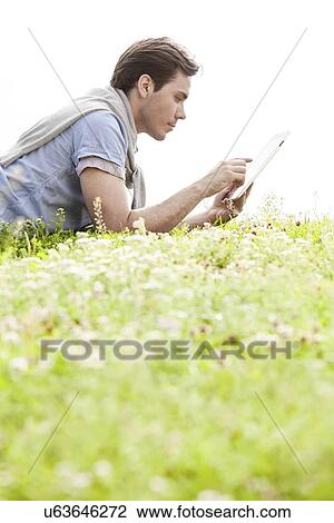 Side view of young man using digital tablet while lying on grass against clear sky View Large Photo Image Stock Image - Side view of young man using digital tablet while lying on grass against clear sky. Fotosearch