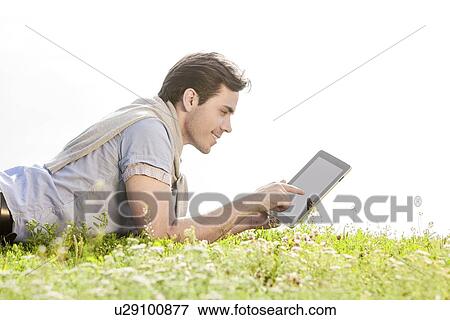 Side view of young man using digital tablet while lying on grass against clear sky View Large Photo Image Stock Photo - Side view of young man using digital tablet while lying on grass against clear sky. Fotosearch