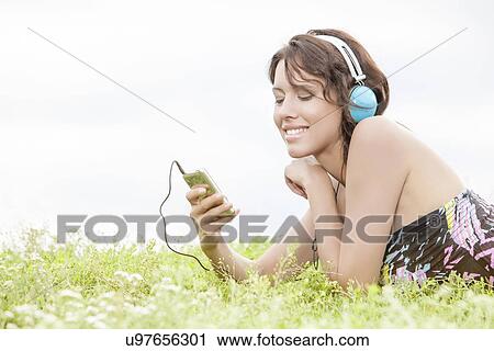 Side view of young woman listening to music through cell phone using headphones while lying on grass against clear sky View Large Photo Image Stock Image - Side view of young woman listening to music through cell phone using headphones while lying on grass against clear sky. Fotosearch