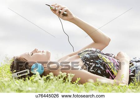 Side view of young woman listening to music through MP3 player while lying on grass against sky View Large Photo Image Stock Photograph - Side view of young woman listening to music through MP3 player while lying on grass against sky. Fotosearch