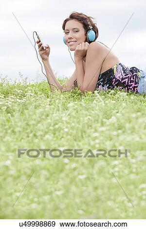 Side view portrait of woman listening to music through cell phone using headphones while lying on grass against sky View Large Photo Image Stock Photo - Side view portrait of woman listening to music through cell phone using headphones while lying on grass against sky. Fotosearch