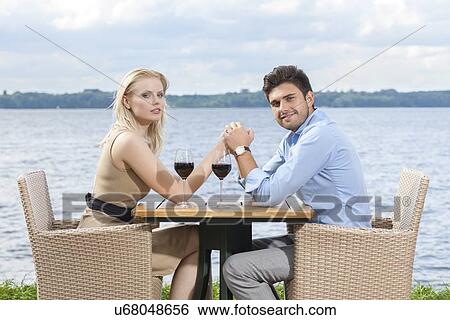 Stock Photograph - Side view portrait of young couple holding hands at outdoor restaurant by lake. Fotosearch
