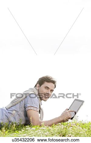 Stock Photograph - Side view portrait of young man using digital tablet while lying on grass against clear sky. Fotosearch