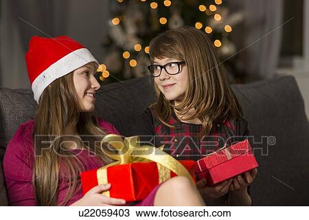 Stock Image - Sisters with Christmas presents looking at each other on sofa. Fotosearch