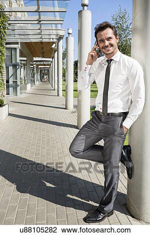 Stock Image - Smiling businessman using mobile phone while leaning on column outside office. Fotosearch