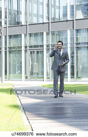 Smiling businessman using mobile phone while walking on path outside office View Large Photo Image Stock Photograph - Smiling businessman using mobile phone while walking on path outside office. Fotosearch