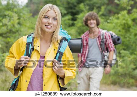 Stock Photo - Smiling female backpacker looking away with man standing in background at forest. Fotosearch