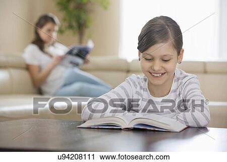 Smiling girl reading book with mother in background at home View Large Photo Image Stock Image - Smiling girl reading book with mother in background at home. Fotosearch