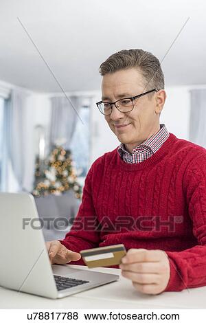 Smiling man using credit card and laptop to shop online at home during Christmas View Large Photo Image Stock Photo - Smiling man using credit card and laptop to shop online at home during Christmas. Fotosearch