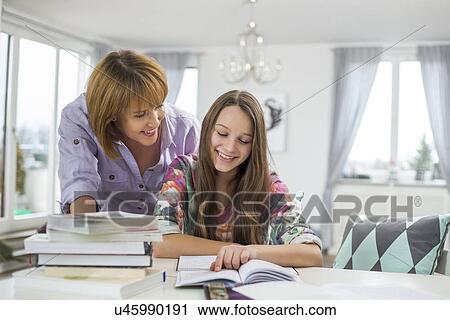 Stock Image - Smiling mother assisting daughter in doing homework at table. Fotosearch
