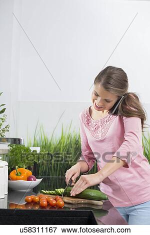 Stock Photo - Smiling woman using cell phone while cutting cucumber at kitchen counter. Fotosearch