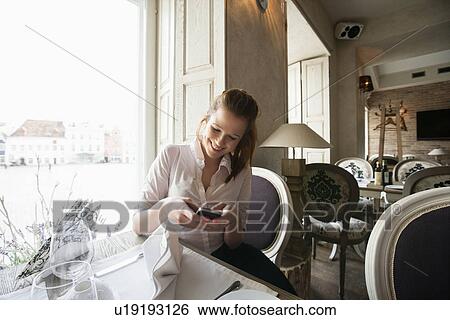 Stock Photograph - Smiling young businesswoman text messaging on cell phone at restaurant table. Fotosearch