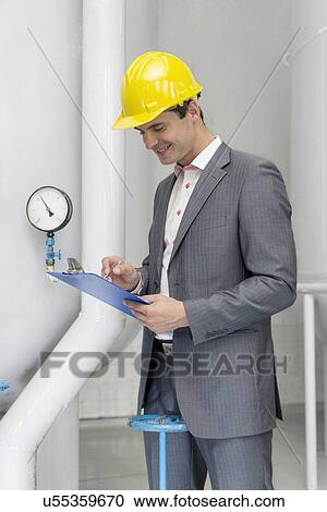 Stock Image - Smiling young male manager writing on clipboard in industry. Fotosearch