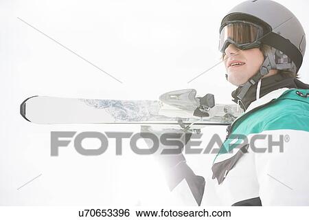 Stock Photograph - Smiling young man carrying skis against clear sky. Fotosearch