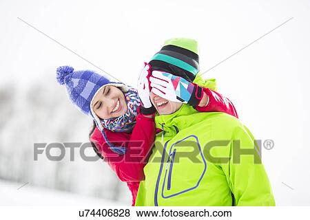Stock Photo - Smiling young woman covering man's eyes in winter. Fotosearch