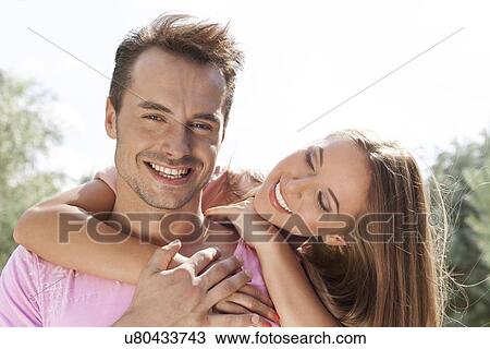 Smiling young woman embracing man from behind in park View Large Photo Image Stock Image - Smiling young woman embracing man from behind in park. Fotosearch