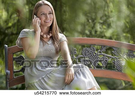 Smiling young woman using mobile phone on park bench View Large Photo Image Stock Image - Smiling young woman using mobile phone on park bench. Fotosearch