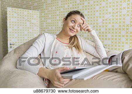 Stock Image - Smiling young woman with books sitting on couch at home. Fotosearch