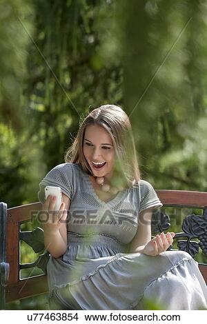 Surprised young woman reading text message on smart phone in park View Large Photo Image Picture - Surprised young woman reading text message on smart phone in park. Fotosearch