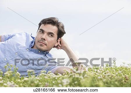 Stock Photograph - Thoughtful man lying on grass against sky. Fotosearch