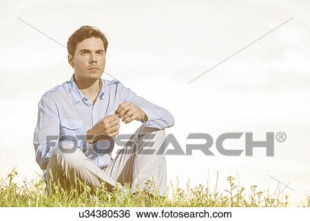 Thoughtful young man looking away while sitting on grass against clear sky View Large Photo Image Stock Photograph - Thoughtful young man looking away while sitting on grass against clear sky. Fotosearch