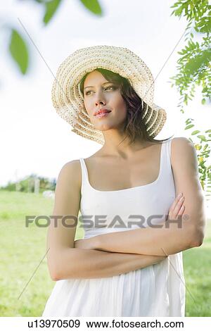 Stock Photo - Thoughtful young woman in sundress and hat standing arms crossed in park. Fotosearch