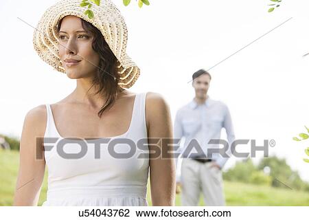Stock Image - Thoughtful young woman looking away with man in background at park. Fotosearch