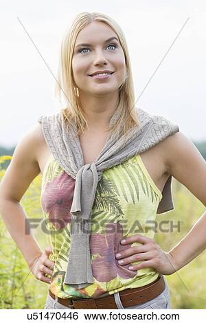 Stock Photograph - Thoughtful young woman looking away while standing with hands on hips in field. Fotosearch