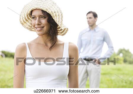 Thoughtful young woman smiling while man standing in background at park View Large Photo Image Stock Image - Thoughtful young woman smiling while man standing in background at park. Fotosearch