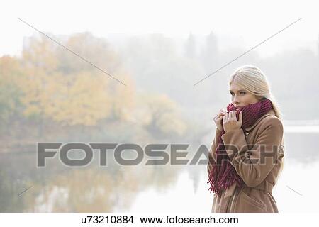 Picture - Thoughtful young woman wearing muffler at lakeside in park. Fotosearch
