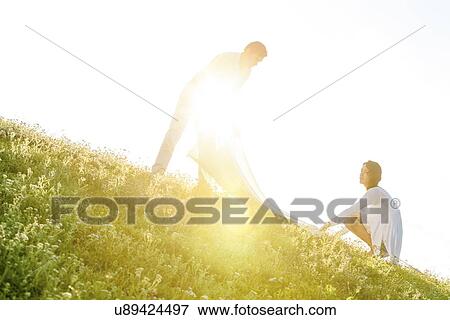 Stock Photo - Tilt image of young couple spreading picnic blanket on grass during sunny day. Fotosearch