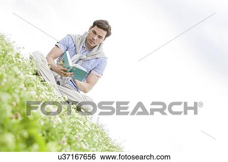 Stock Photograph - Tilt image of young man reading book while sitting on grass against sky. Fotosearch