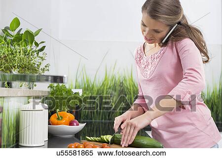 Stock Photography - Woman using cell phone while cutting cucumber at kitchen counter. Fotosearch
