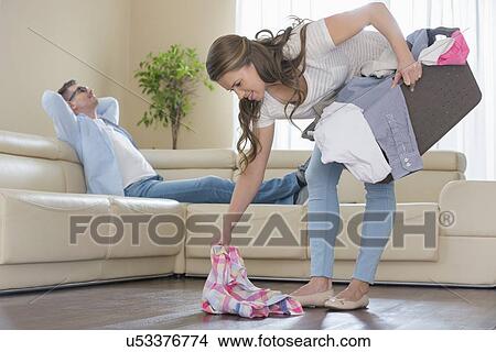 Picture - Woman with laundry basket picking clothes while man relaxing on sofa in background. Fotosearch