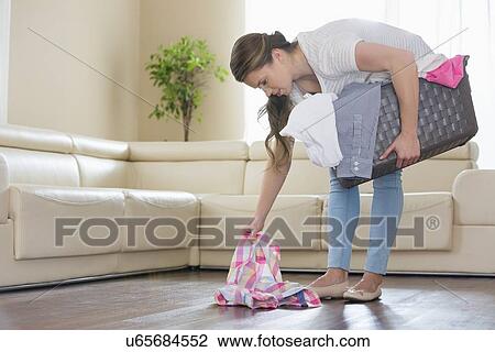 Stock Image - Woman with laundry basket picking clothes from floor in living room. Fotosearch
