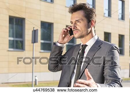 Young businessman conversing on cell phone against office building View Large Photo Image Stock Photo - Young businessman conversing on cell phone against office building. Fotosearch